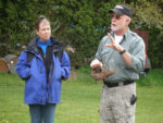 Farmer talking out to a group of people holding a root with a woman to his left looking at him