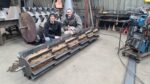 two men kneeling and posing for a photo in front of a roller in a machine shop