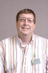 A headshot of a man with orange-brown hair with glasses and a striped collared shirt.