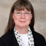 Deborah Cavanaugh-Grant headshot with brown hair and bangs wearing a black cardigan and a spotted scarf