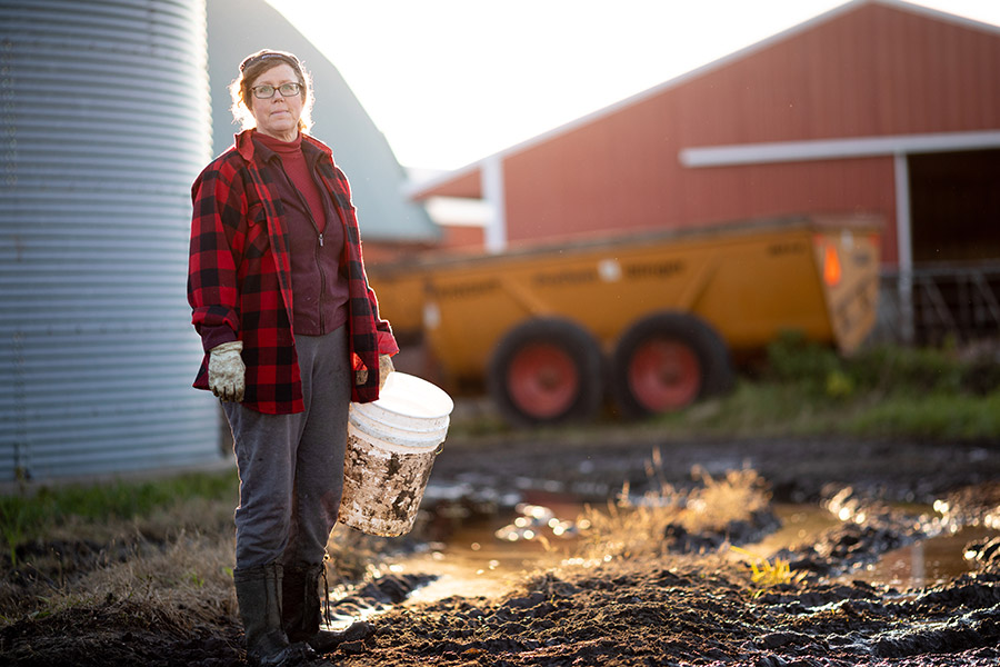 Woman in red coat standing in front of a barn with a white 5 gallon bucket in her hand.