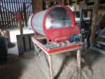A red cylindrical drum with screened walls sits on a table above a pile of dust.