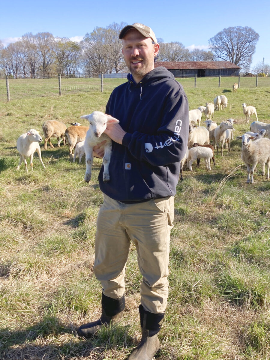a man in a field holding a baby sheep wtih a small herd behind him