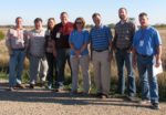 A group of men and woman standing together, smiling for a picture in front of a field.