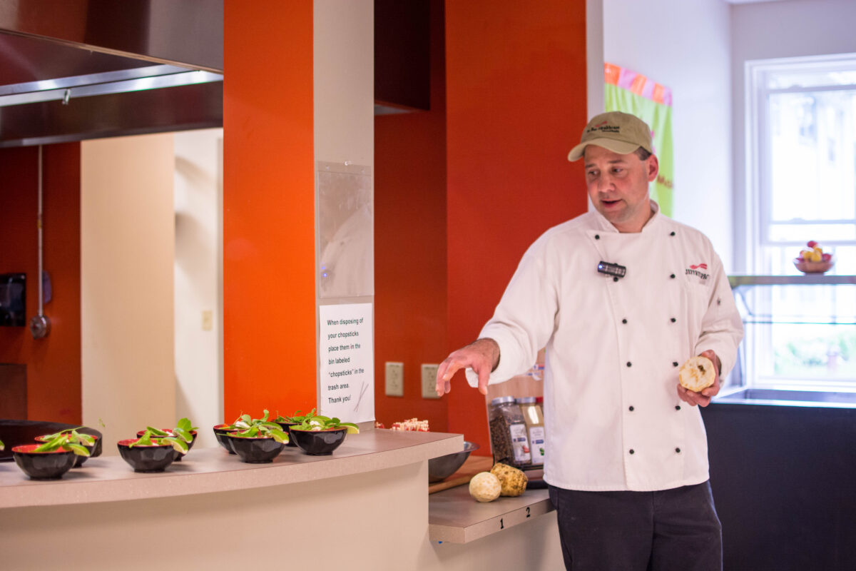 A person in a white coat pointing to prepared salad bowls on a counter in an orange kitchen