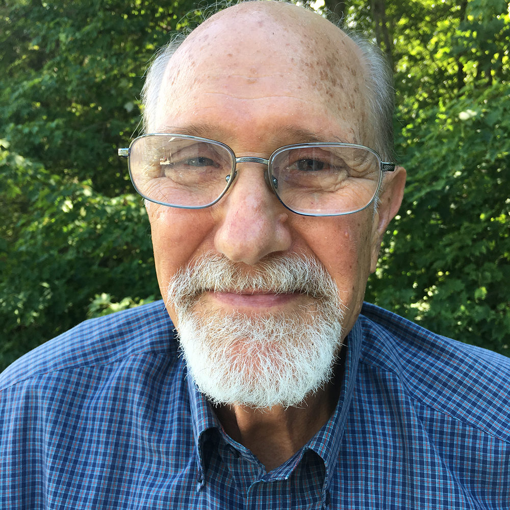Headshot of a man without hair and big glasses and a goatee with a blue plaid shirt