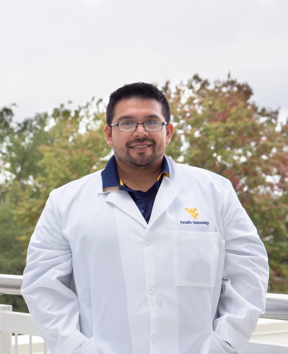 A man with dark hair, a white lab coat, and glasses smiling for a headshot outside