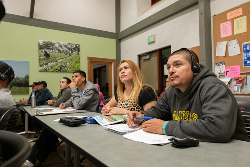Farmers in a classroom during a workshop, one is wearing a headset that provides the discussion in Spanish