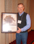 Jim Stordahl holding up a framed picture of a tree wearing a name tag