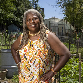 A female Black farmer wearing a yellow dress poses at an urban farm