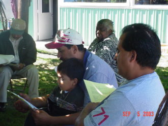 A man reads to a child on his lap, with other people sitting around in chairs outside