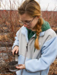 A young woman holding a blade to a metal pot of soil in the woods.