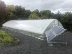 A high tunnel with a solar panel array in front, a wind turbine behind, and fresh gravel underneath from installing geothermal pipes