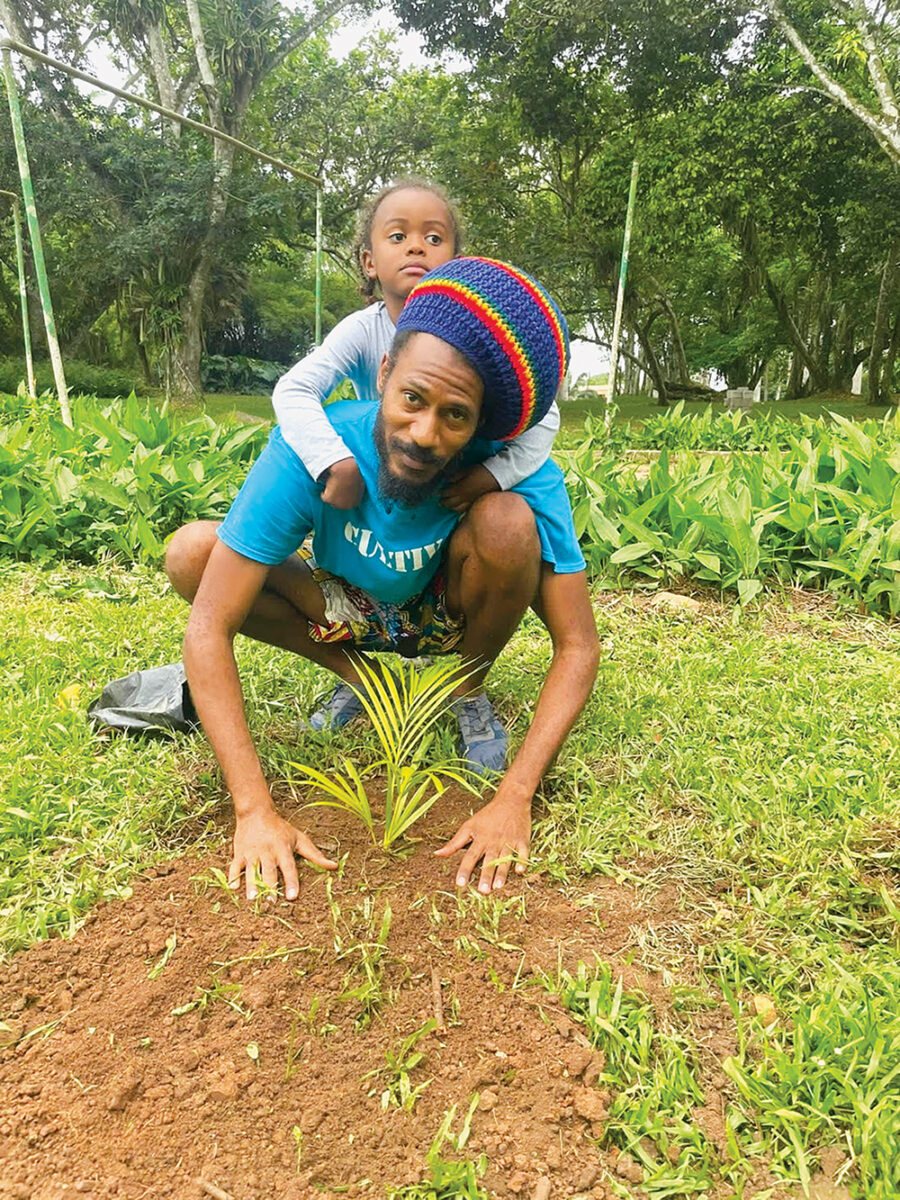 A daughter climes on top of their father while he kneels down in the field over a plant