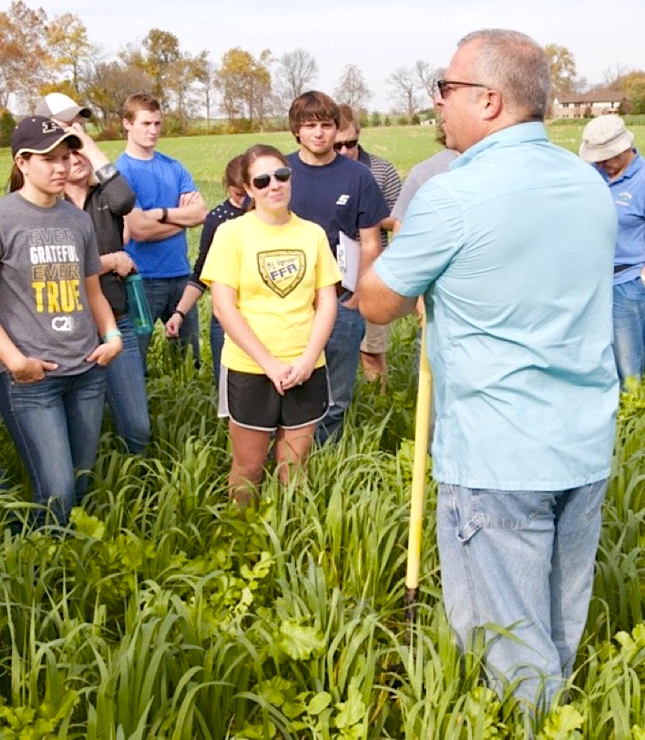 Dan DeSutter is standing in a field of a diverse mix of knee-high cover crops while talking with a group of young adults.