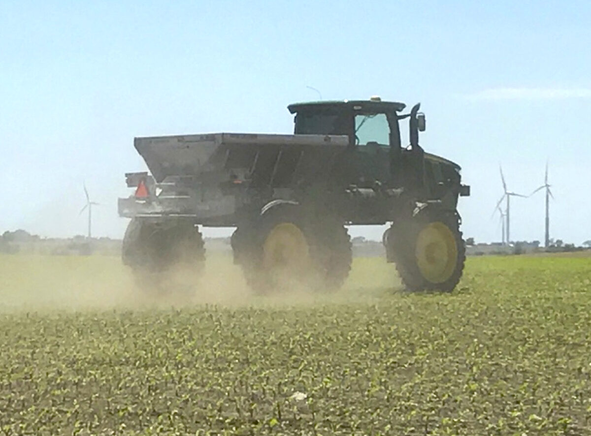 A high-clearance seeder, with very tall tires, is driving through a field that appears to have crop stubble and maybe small corn plants.