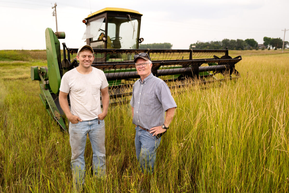 two farmers standing in a field of tall oats with a combine behind them