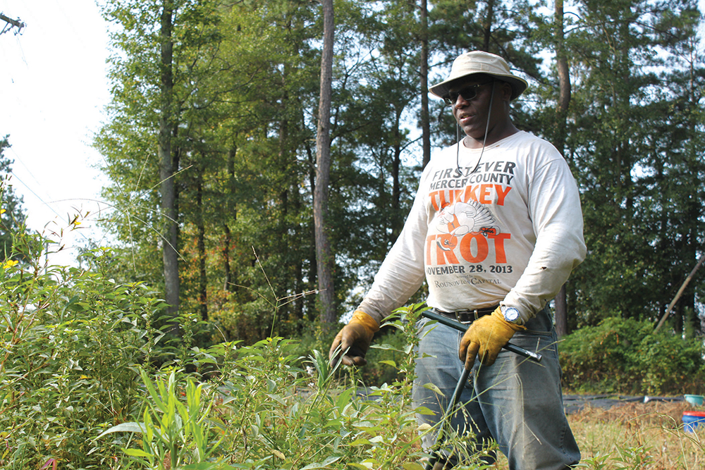 Man working in the field with a hat, sunglasses, long sleeve shirt, and gloves, holding tools