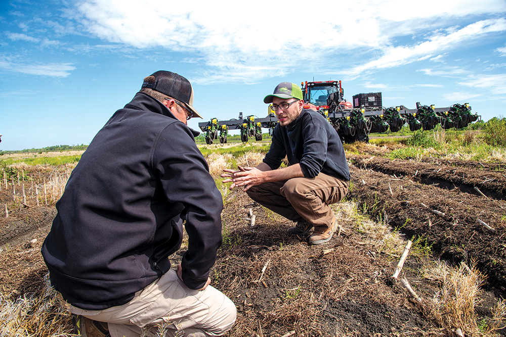 Two people kneeling in a farming field, in the brown dirt, with a no-till planter behind them