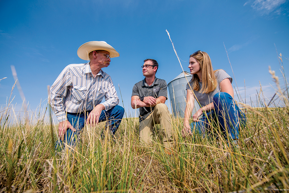 Three people - two women and one man - kneeling and talking on a grassy rangeland