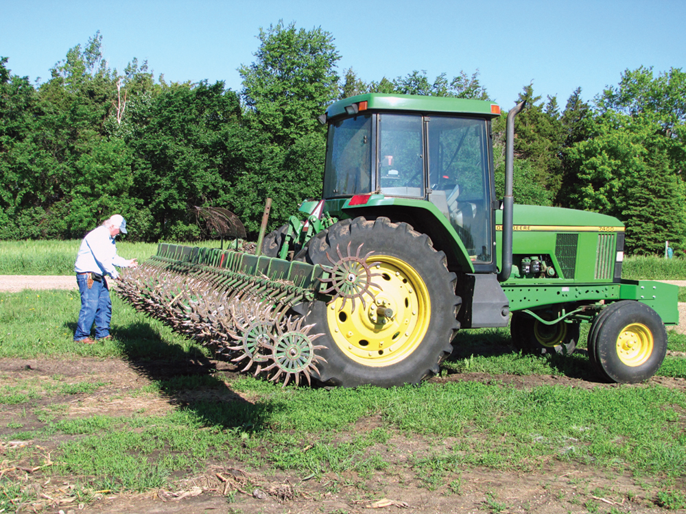 A farmer inspecting the wheels of a rotary hoe mounted onto the back of a tractor.