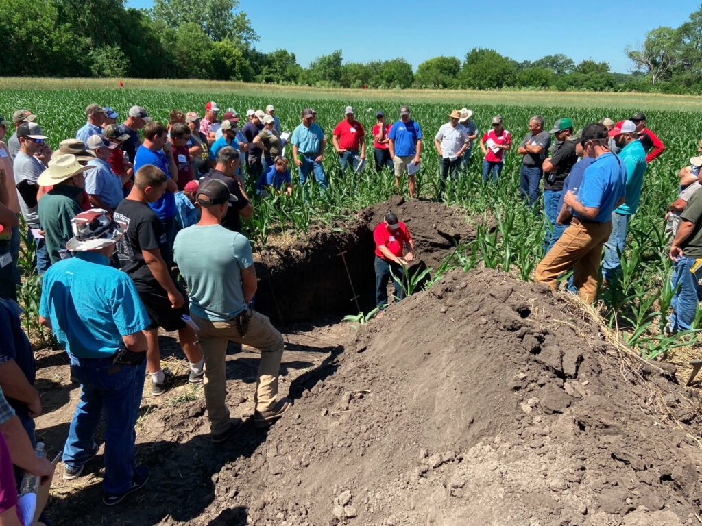 A man stands in a soil pit and is surrounded by visitors at a field day