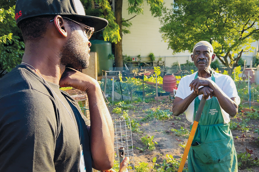 A young farmer looks back to talk to an older farmer holding a tool in the middle of a garden