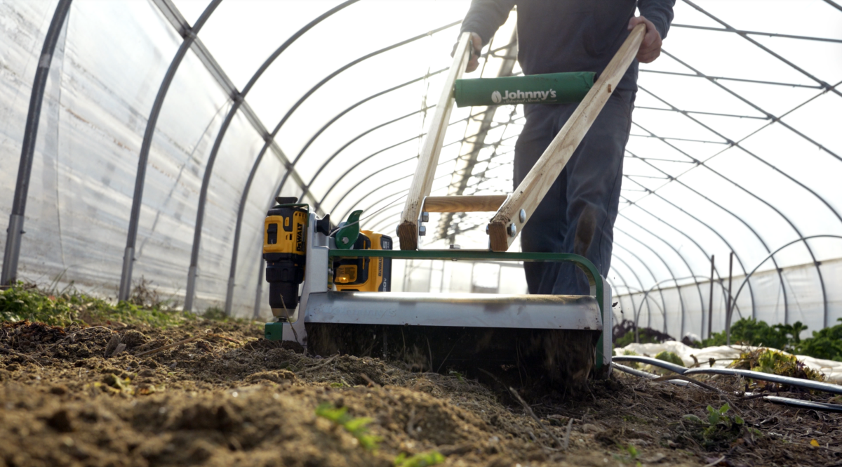 A person is operating a tilther over a bed inside a high tunnel.
