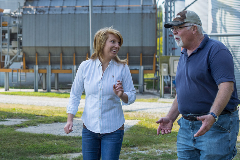 A woman is chatting with her father at their farm, with large grain storage buildings behind them