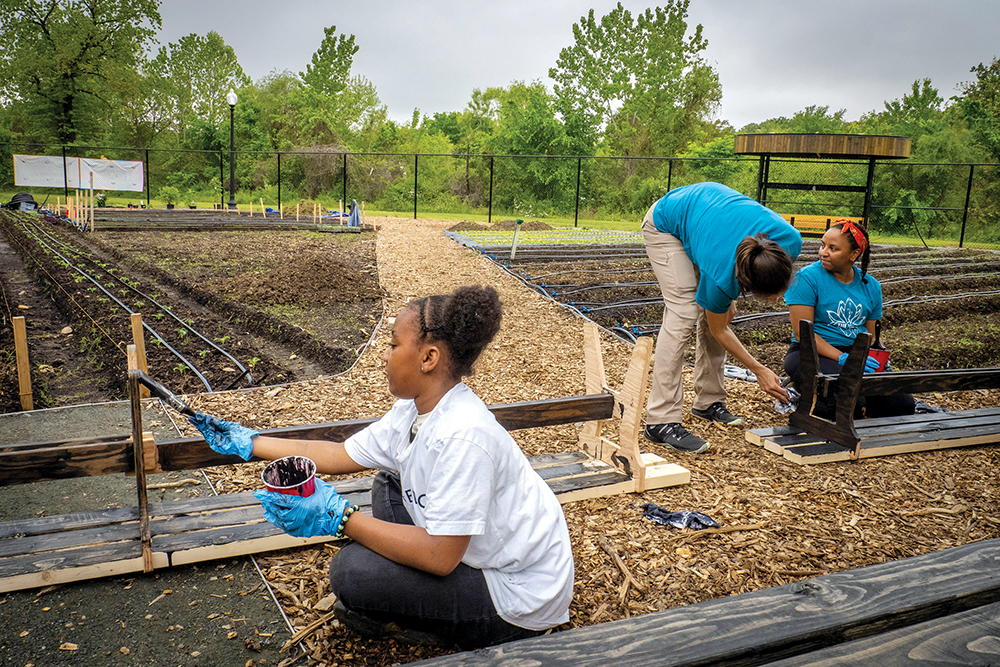 Two kids working under the guidance of an adult in urban beds, painting wooden bed posts